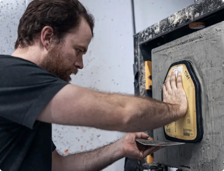 Man pressing a hand against a yellow sanding tool fixed to a wall, smoothing a gray surface.