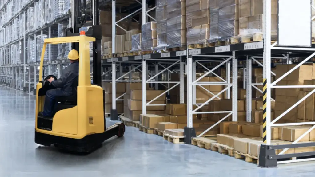 Worker operating a yellow forklift driving down an aisle in a warehouse with shelves stacked with cardboard boxes.
