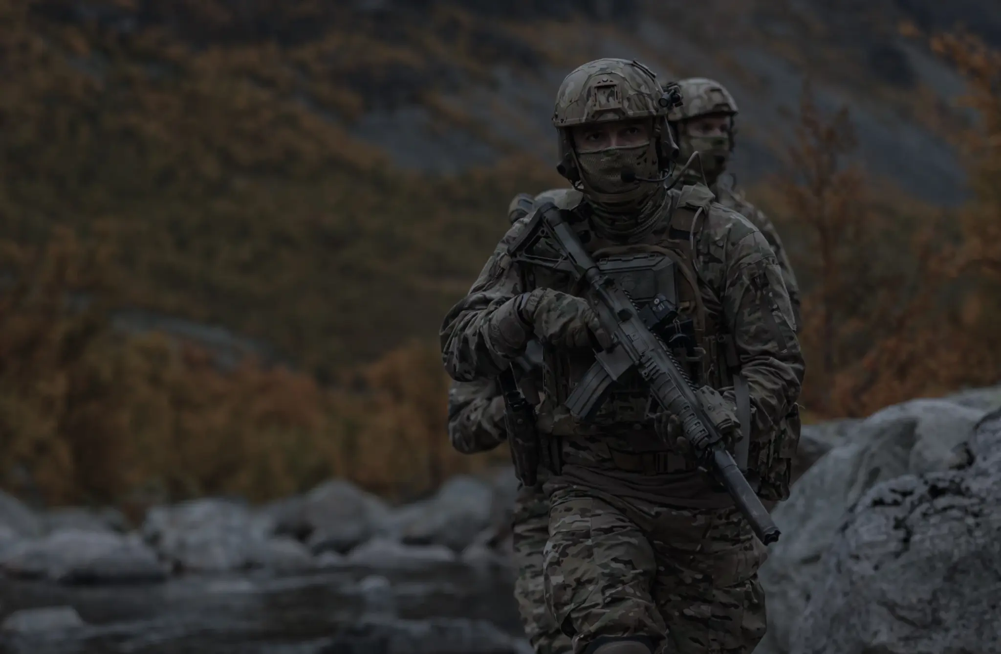 Two soldiers in camouflage gear with rifles walking near a rocky riverbank in a forested mountainous area.