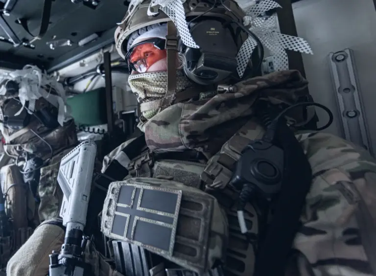 Norwegian soldiers in camouflage uniforms and tactical gear inside a military vehicle, one prominently wearing a helmet and face covering with clear goggles.