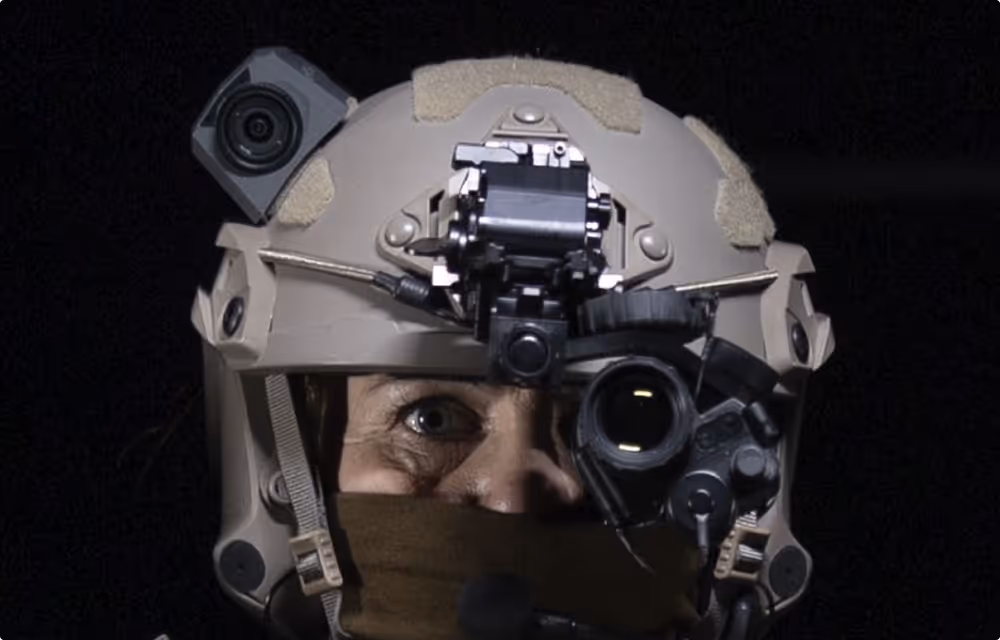 Person wearing a tan tactical helmet equipped with a night vision device and a mounted camera against a black background.