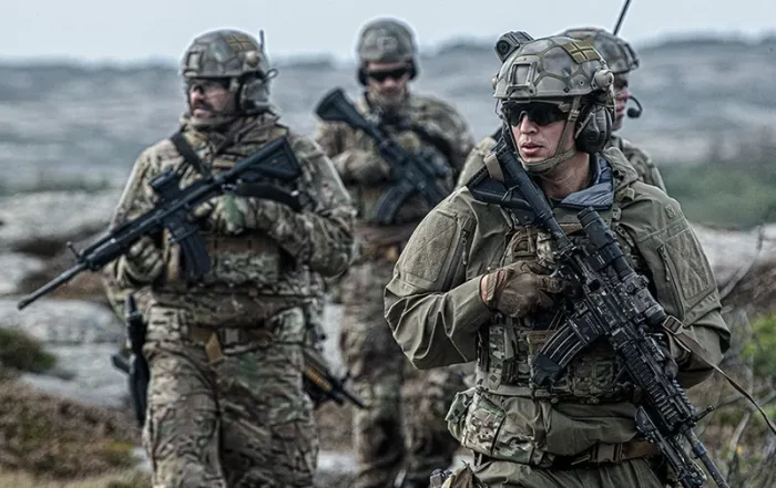 Four soldiers in camouflage uniforms and helmets carrying rifles, walking outdoors in a rocky terrain.