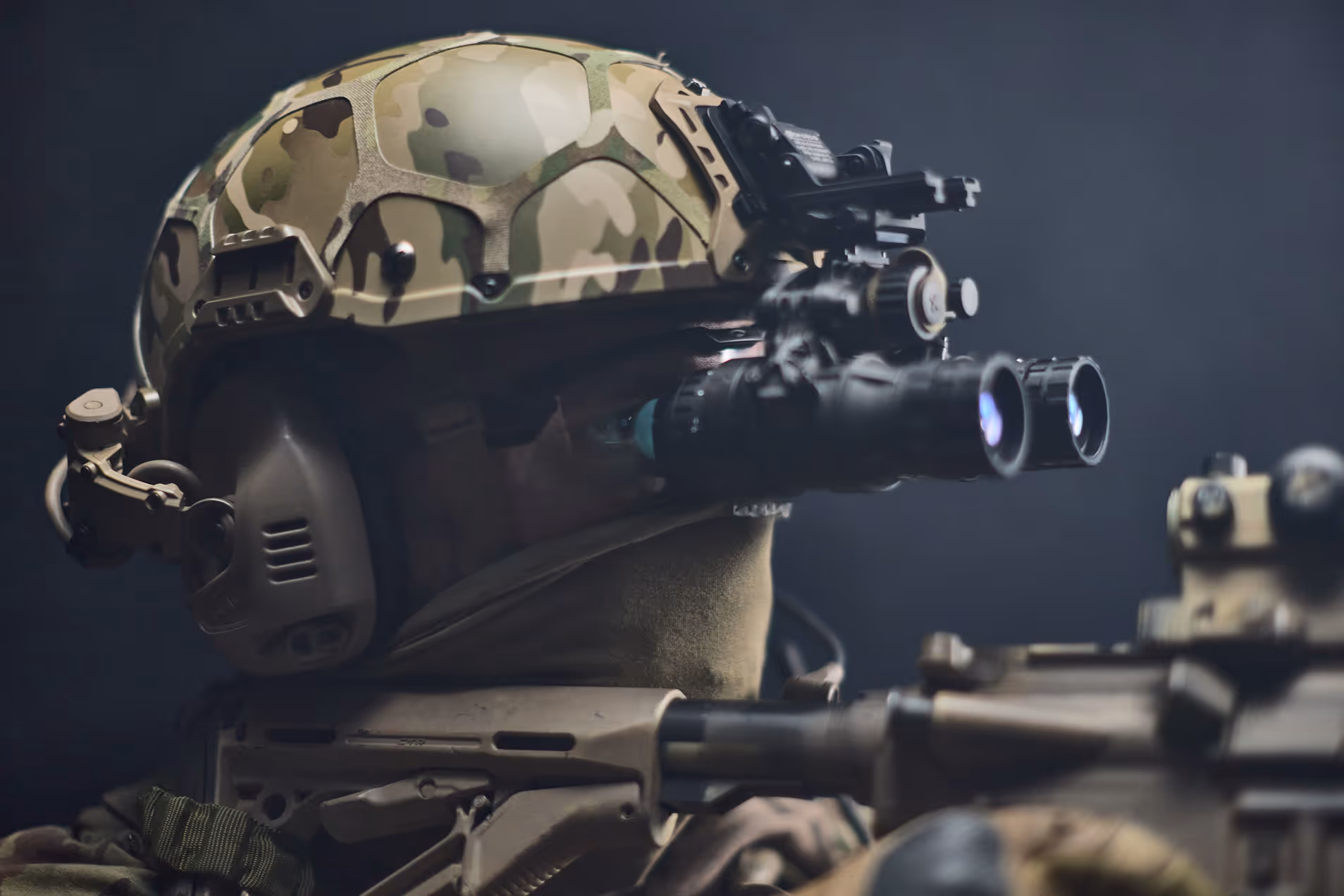Soldier wearing camouflage helmet equipped with night vision goggles aiming a rifle.