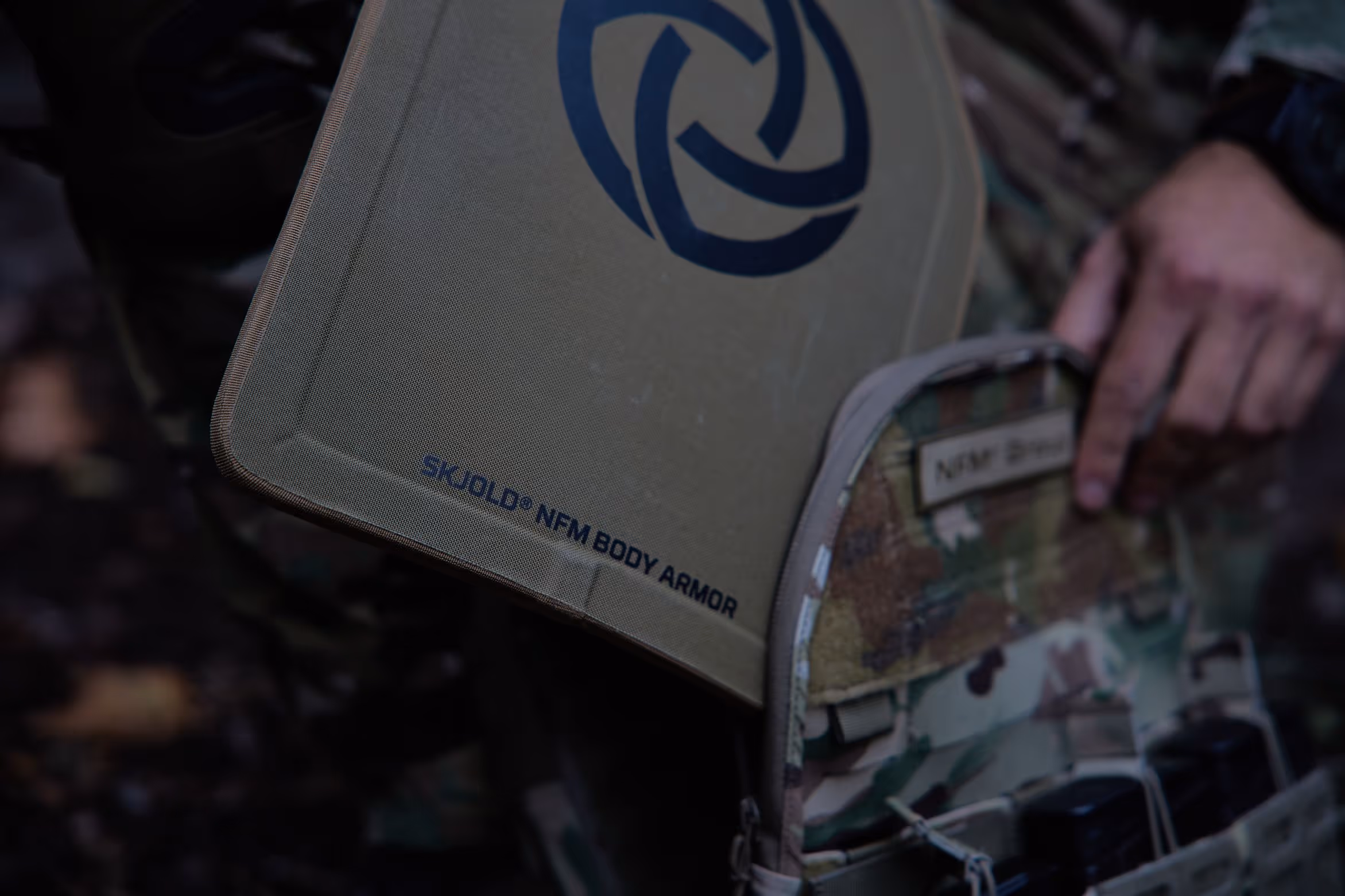 Person holding a tan SKJOLD NFM body armor plate near a camouflage tactical vest.