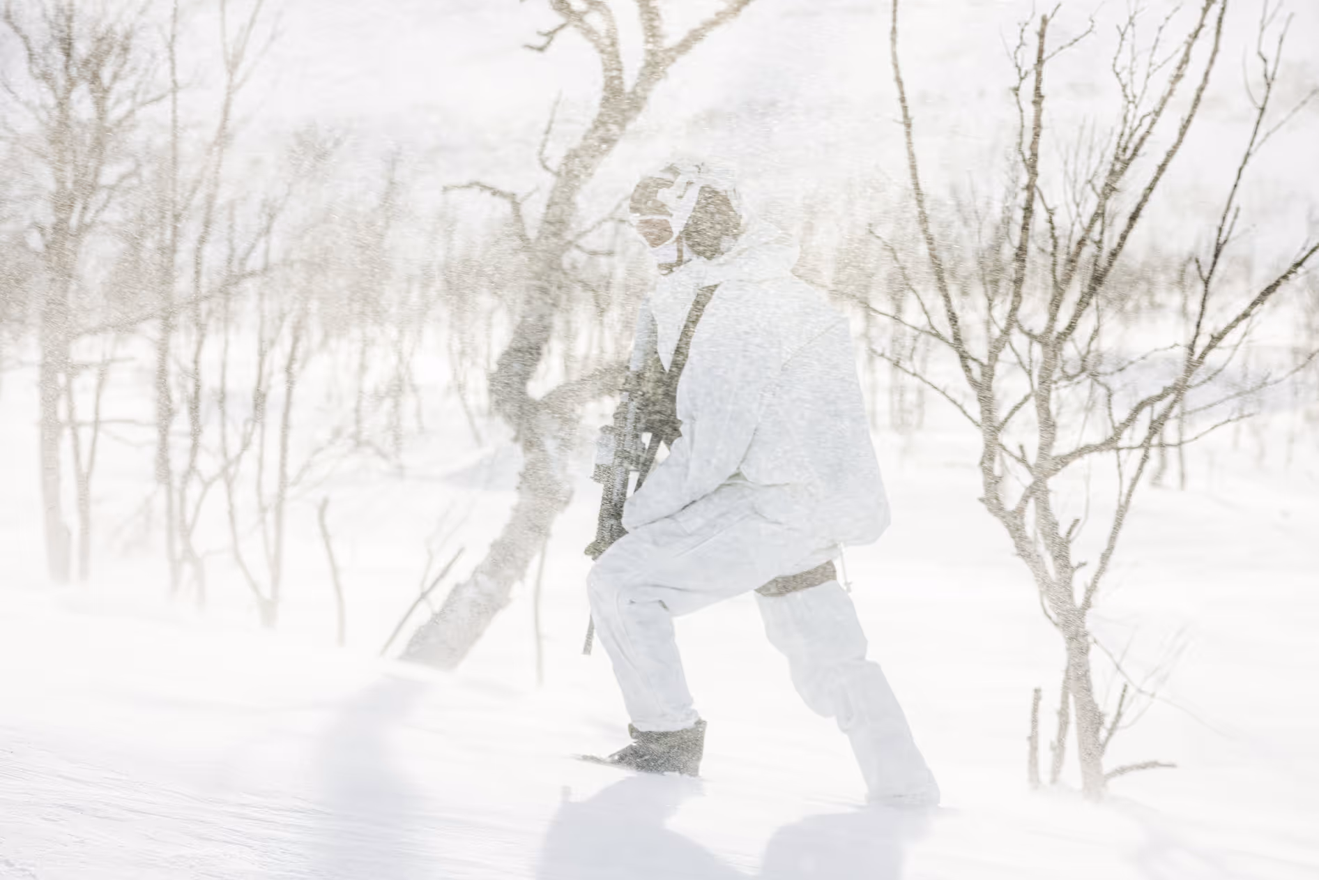Soldier dressed in white camouflage gear walking through snowy terrain with bare trees and falling snow.