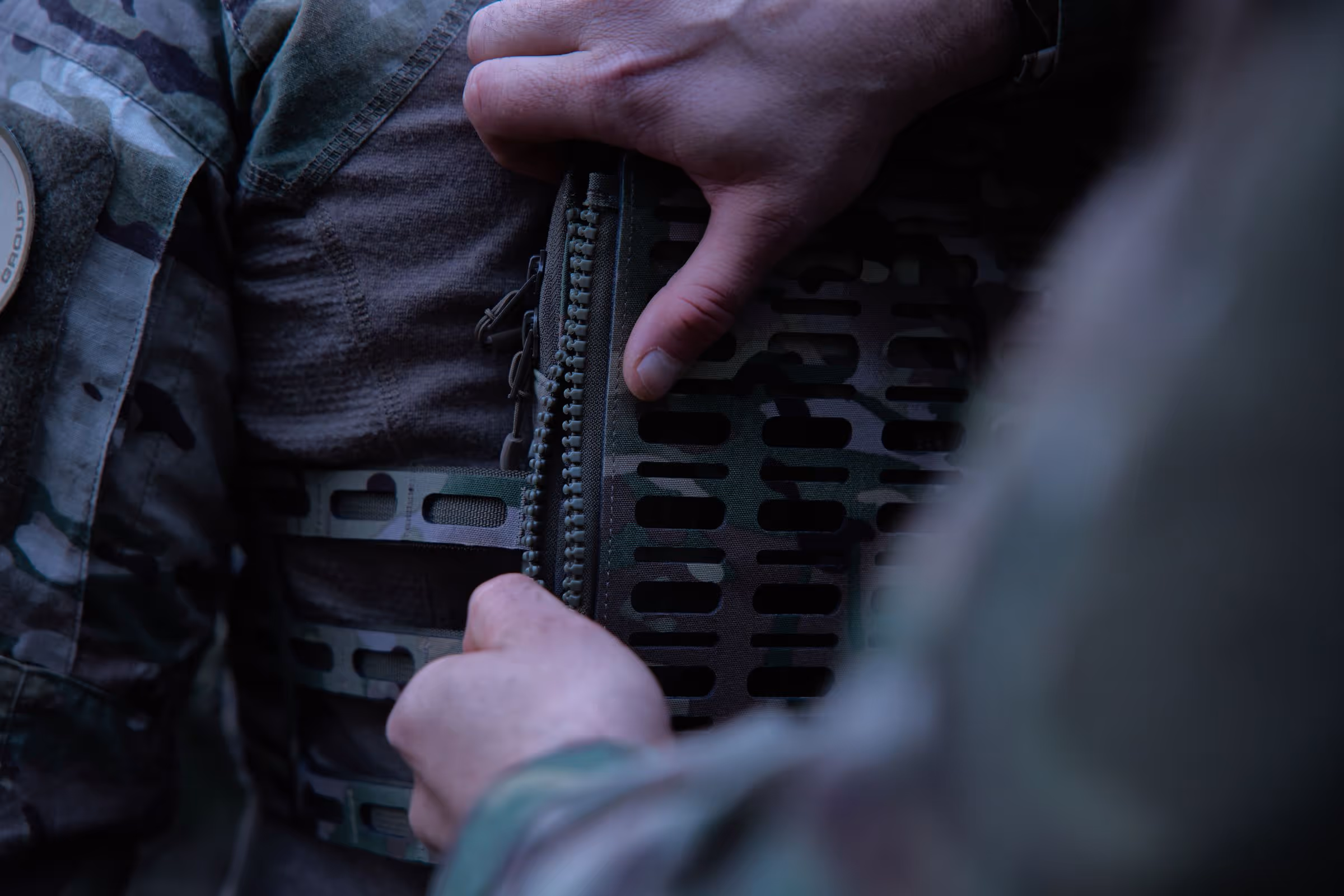Close-up of two hands fastening a zipper on camouflage tactical gear worn over a brown shirt.