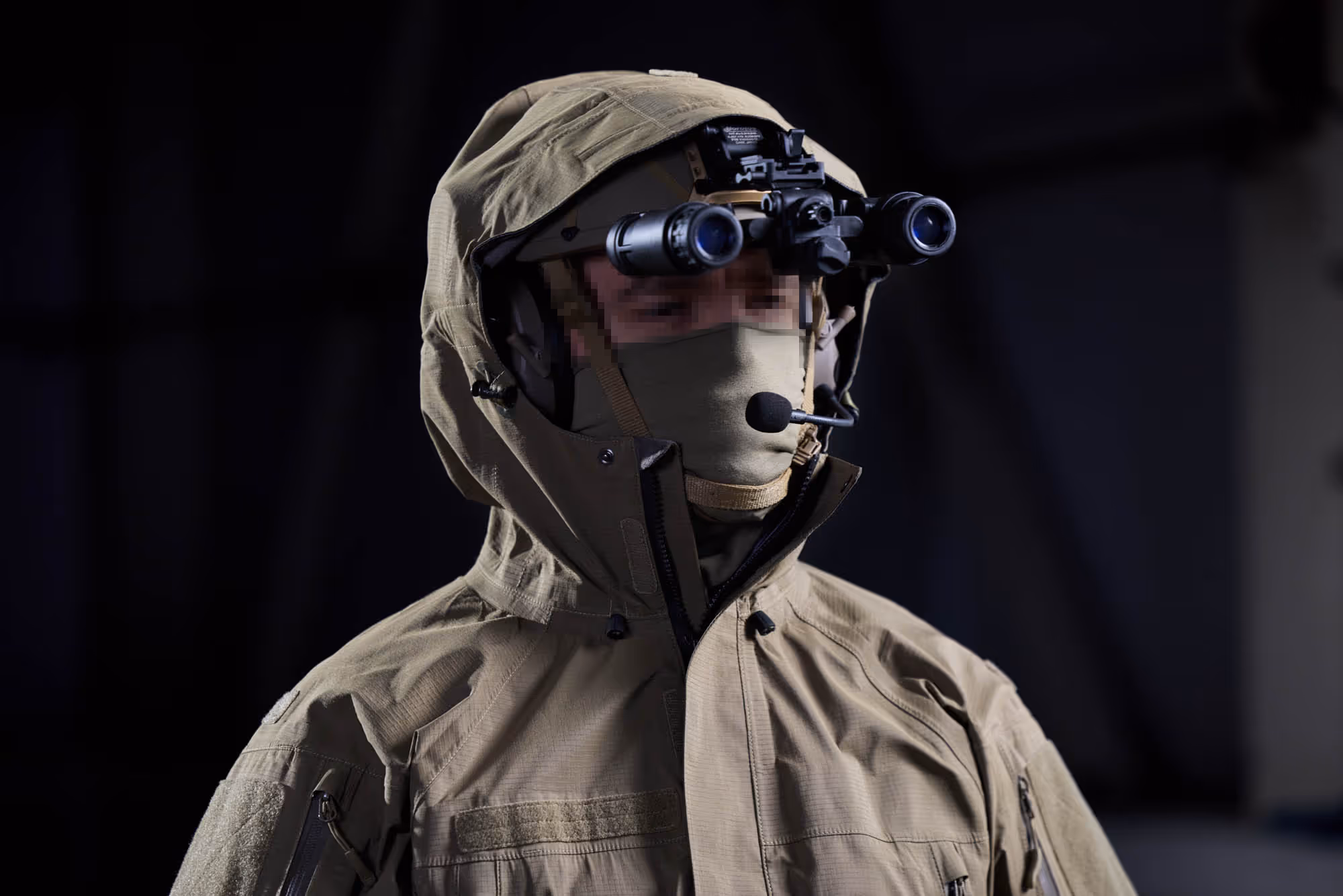 Person wearing military tactical gear including night vision goggles, a headset microphone, and a tan hooded jacket with a face mask.