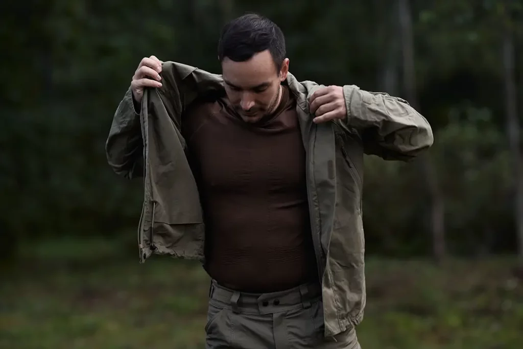 Man outdoors removing an olive-green jacket to reveal a brown fitted shirt and matching pants.