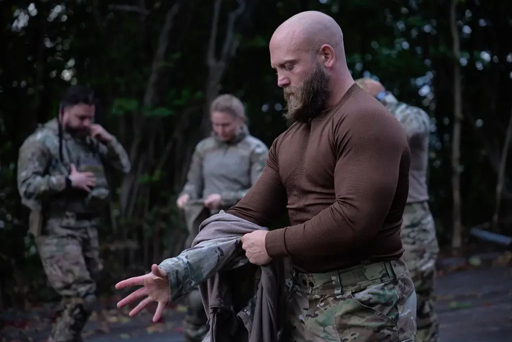 Bearded man in brown shirt putting on a camouflage jacket outdoors with other people in military gear in the background.