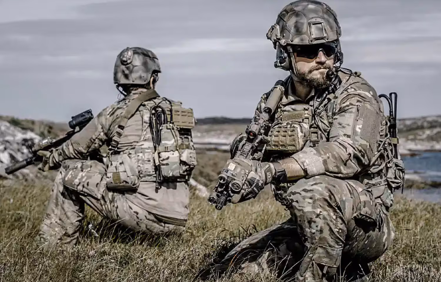 Two soldiers in full camouflage gear and helmets crouching in grassy terrain, holding rifles with tactical attachments.