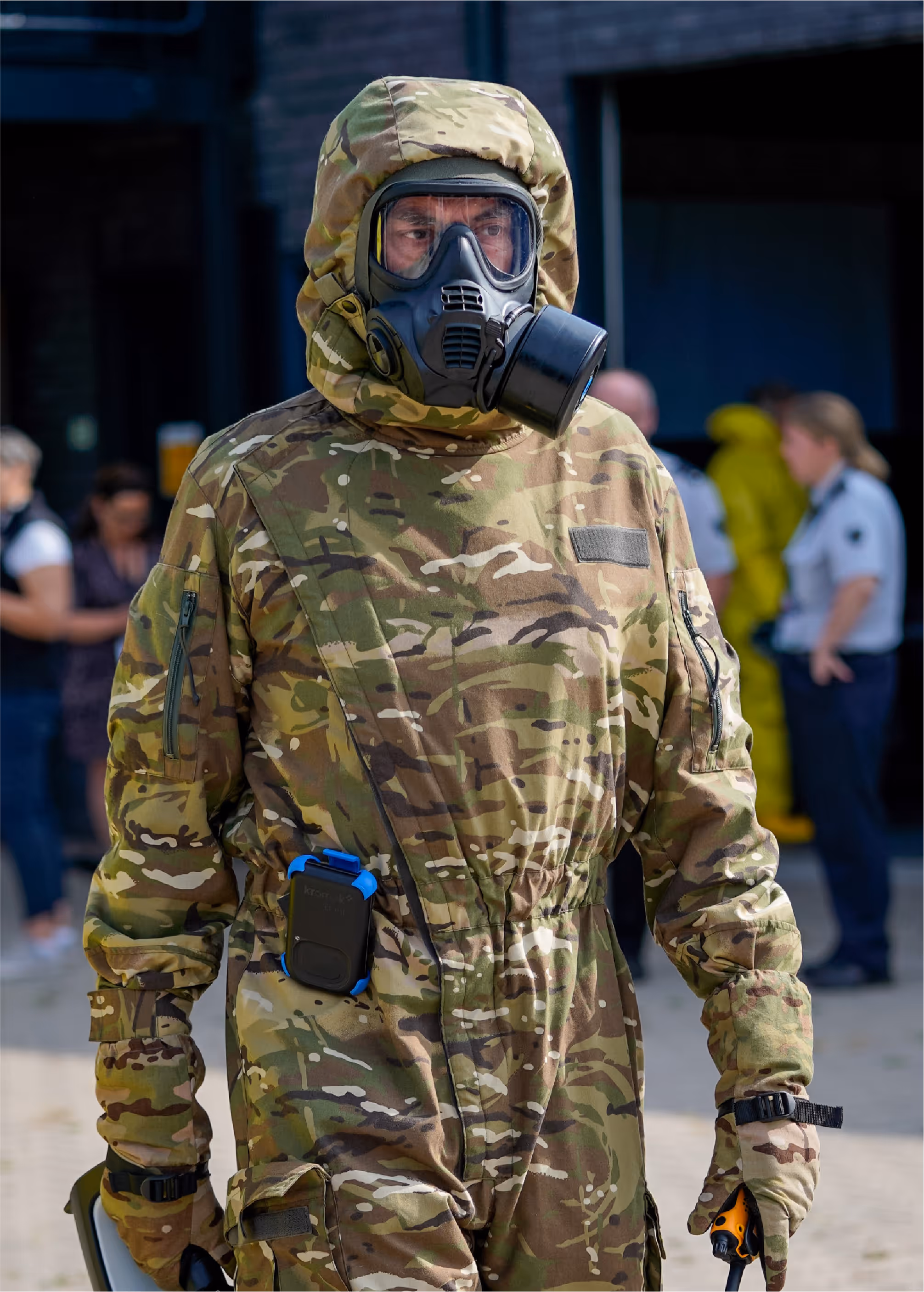 Person wearing camouflage protective suit and a black gas mask with clear eye lenses.