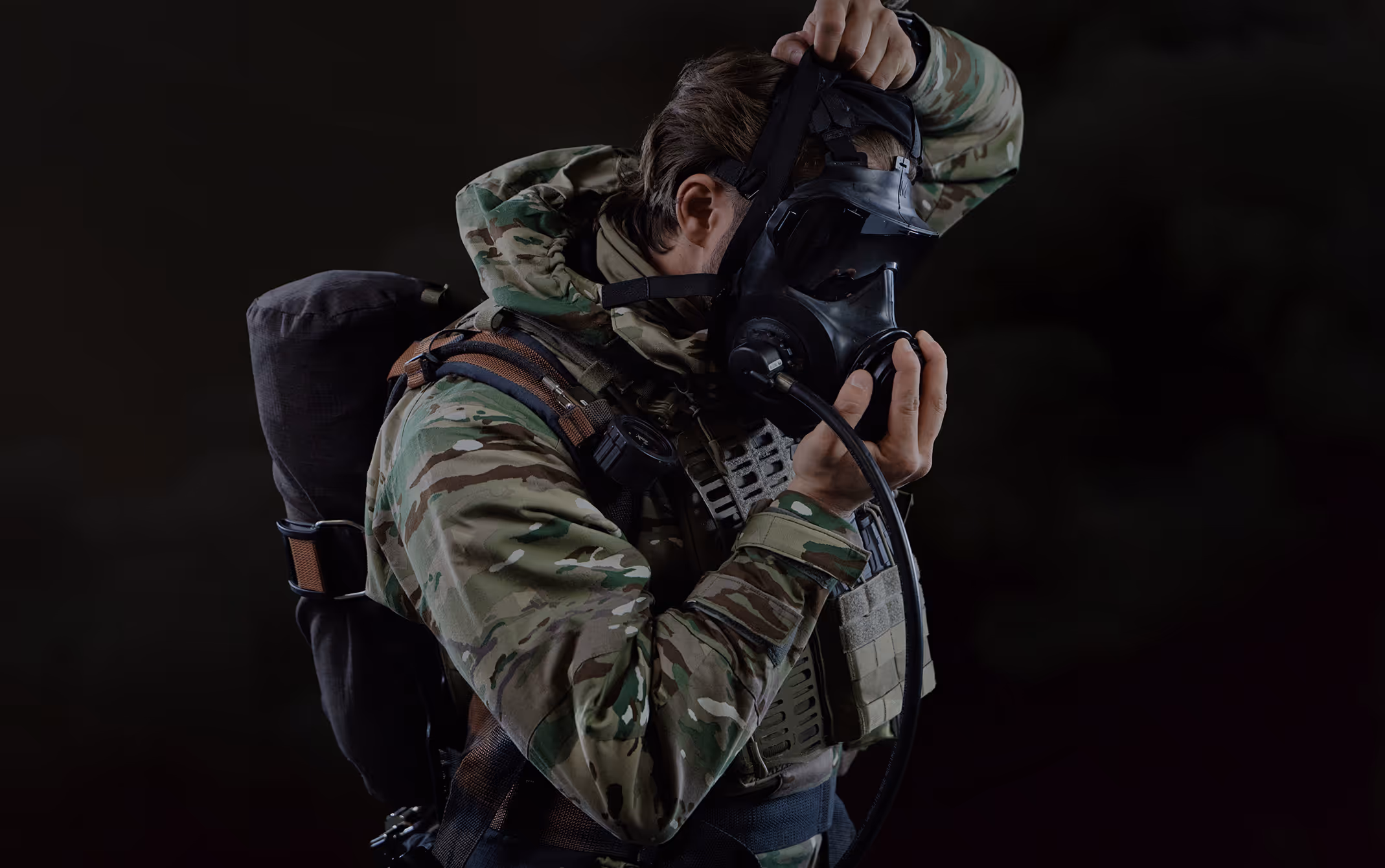 Soldier in camouflage uniform putting on a black gas mask against a dark background.