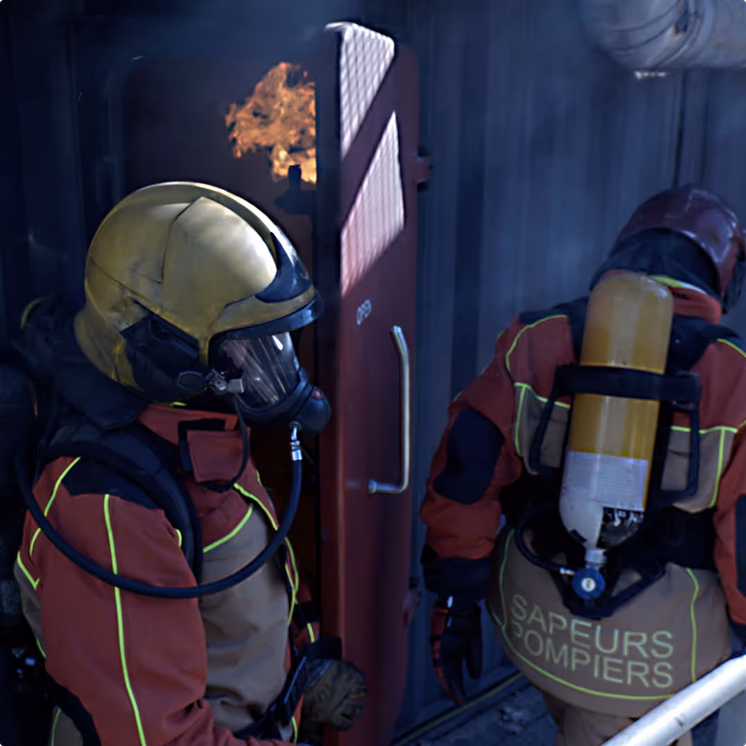 Two firefighters in full gear, one with a yellow helmet and both wearing oxygen tanks, standing near a door with fire visible inside.
