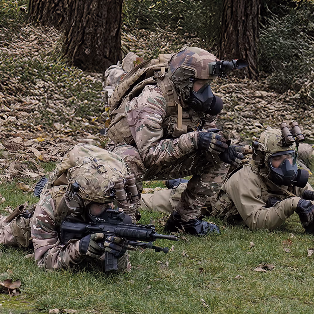 Three soldiers in camouflage gear and gas masks positioned on grass near trees, with one soldier aiming a rifle while others appear to be coordinating.