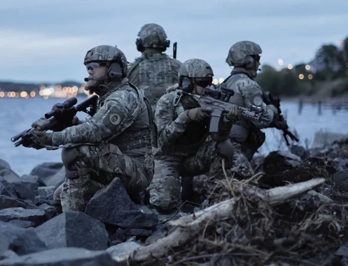 Four soldiers in camouflage gear and helmets holding rifles while positioned on rocky terrain near water at dusk.