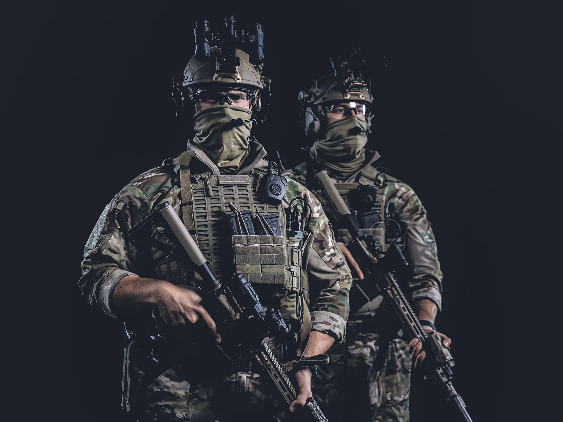 Two soldiers in camouflage gear wearing helmets with night vision goggles and face coverings, holding rifles against a dark background.