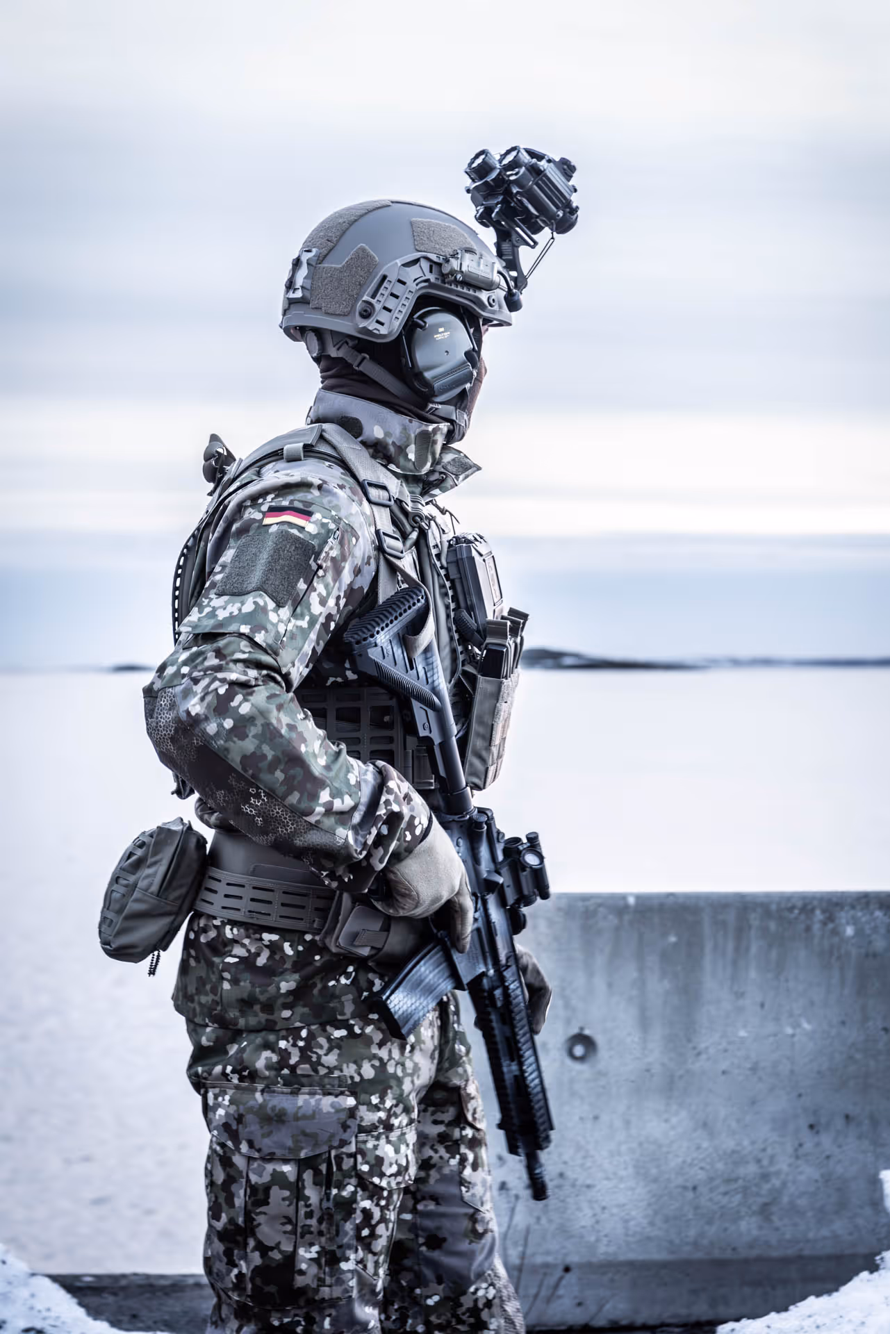 Soldier in camouflage uniform and tactical gear holding a rifle, standing near a concrete barrier with a snowy landscape in the background.
