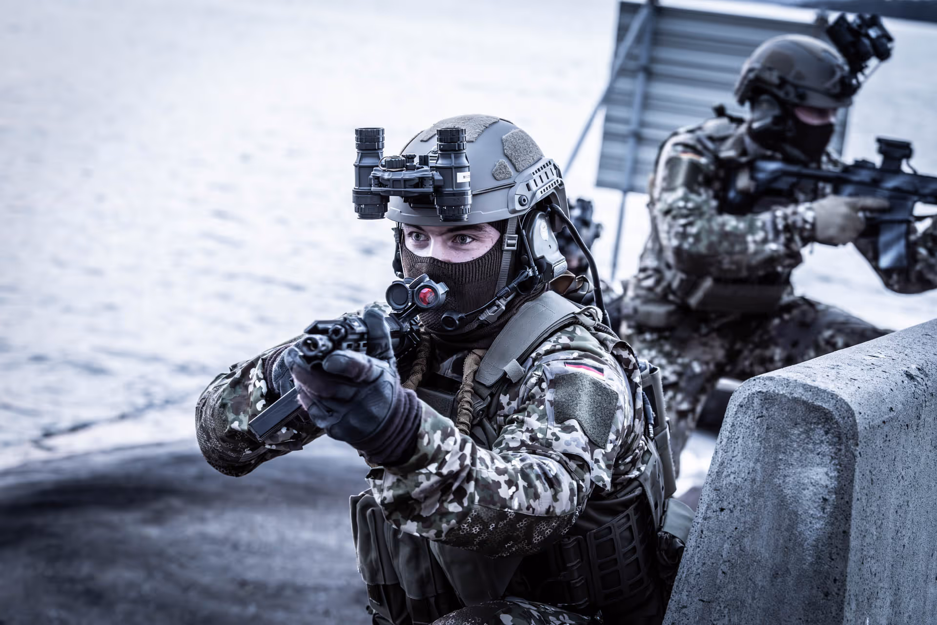 Two soldiers in camouflage gear and helmets aiming rifles near a waterfront.