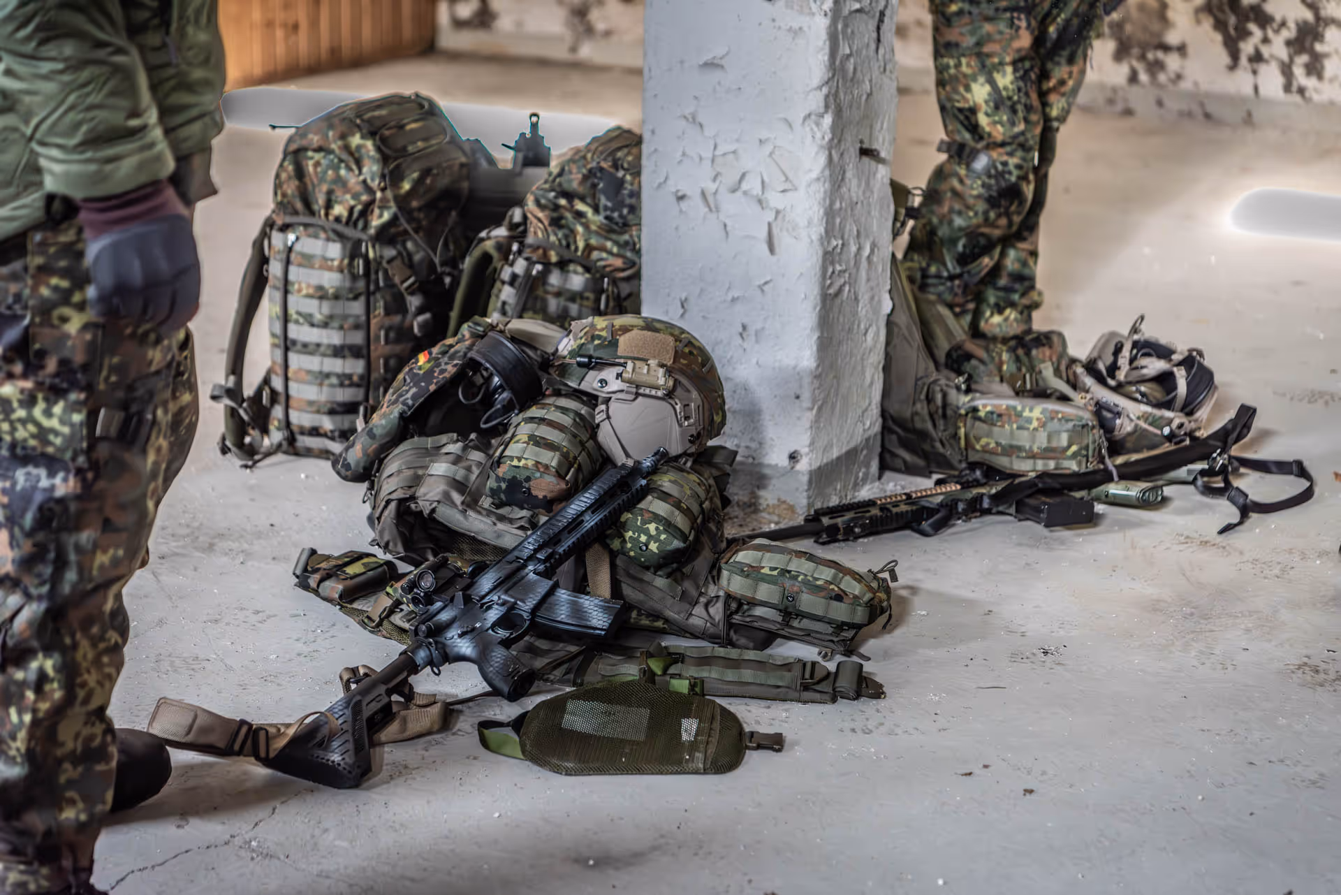 Military gear including rifles, helmets, backpacks, and tactical vests arranged on the floor around a concrete pillar with soldiers in camouflage standing nearby.