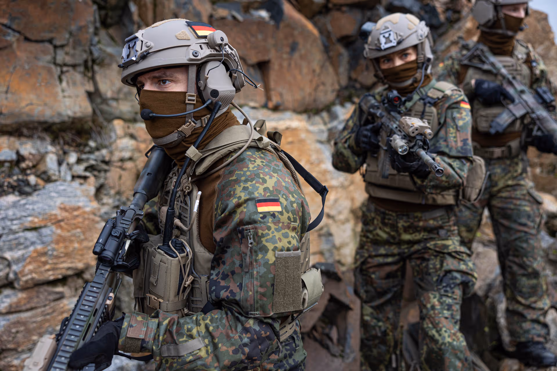 Three soldiers in German camo uniforms and helmets with German flag patches, holding rifles and positioned near rocky terrain.