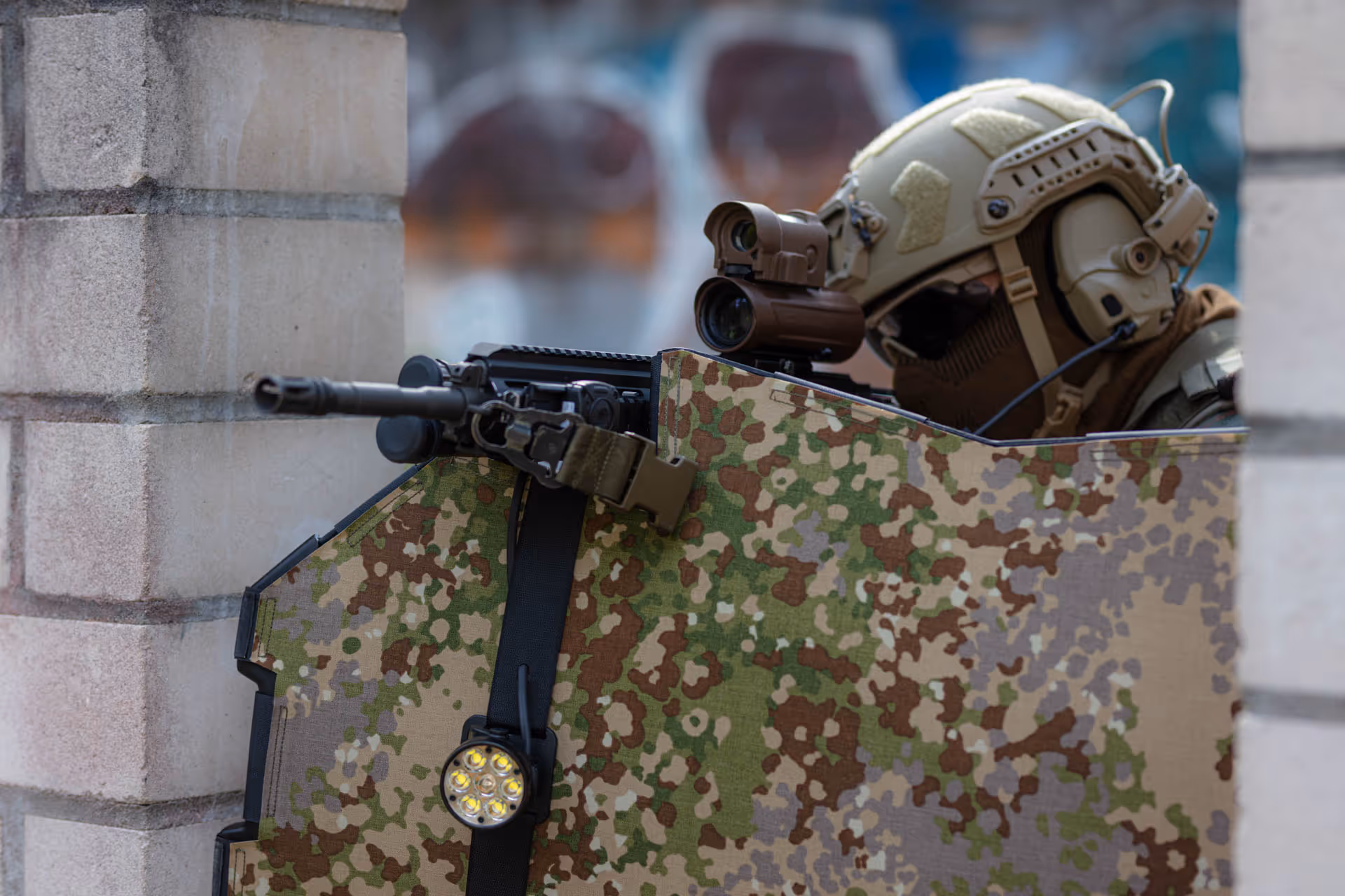 Soldier in tactical gear aiming rifle behind a camouflage ballistic shield near a concrete wall.