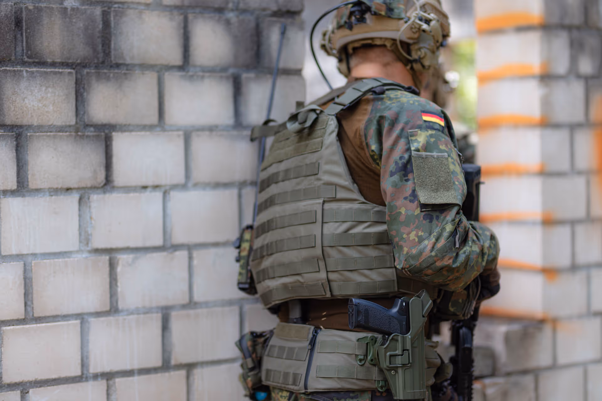 Soldier in camouflage uniform and tactical vest standing with back to the camera near a brick wall.