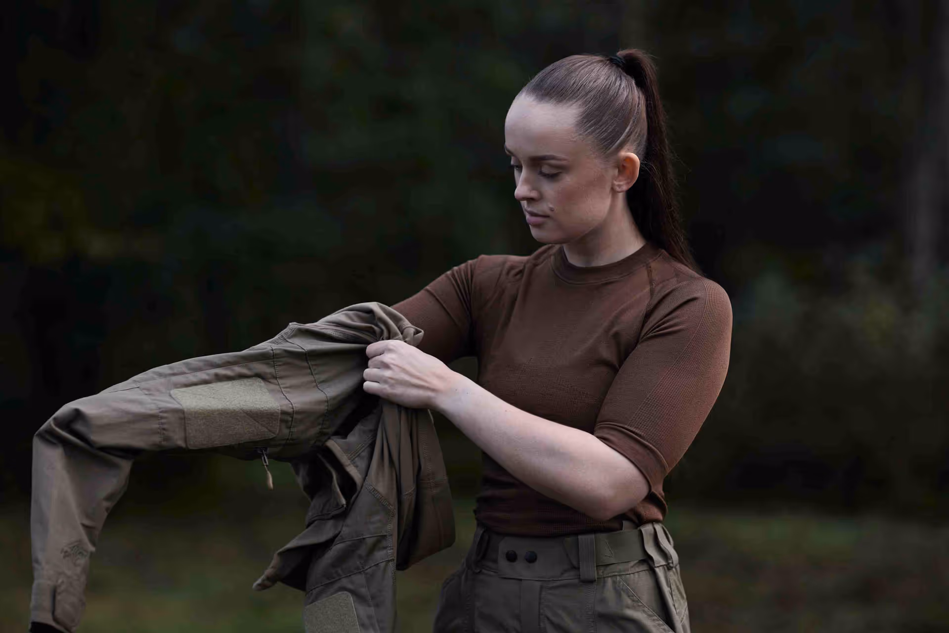 Woman with a ponytail wearing a brown top and green pants holding a green jacket outdoors.