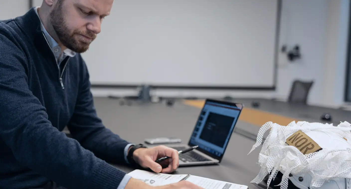 Man sitting at a conference table, writing in a notebook with a laptop and a safety helmet nearby.