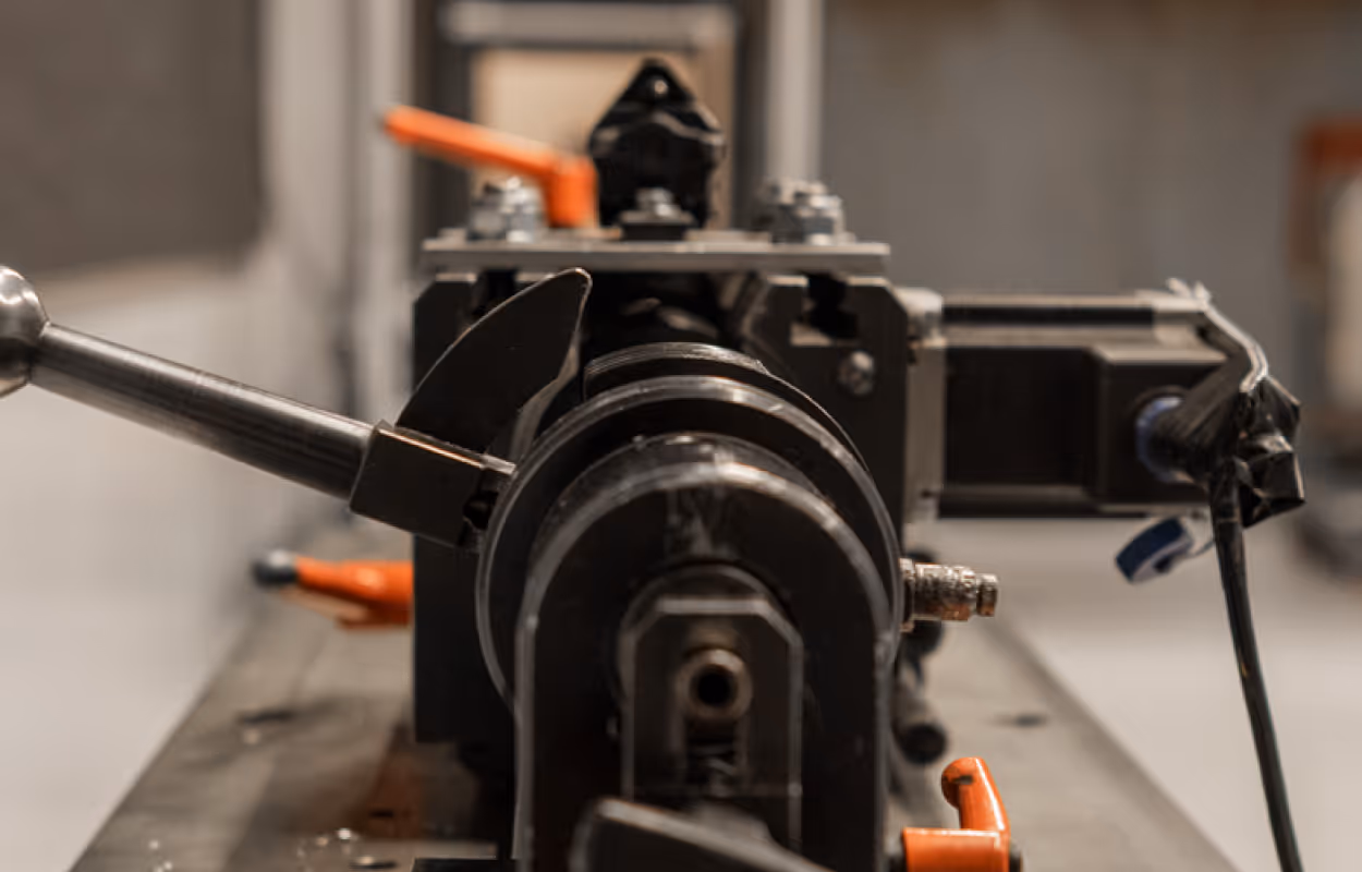 Close-up of a metal machining tool with an attached handle and orange clamps on a workbench.