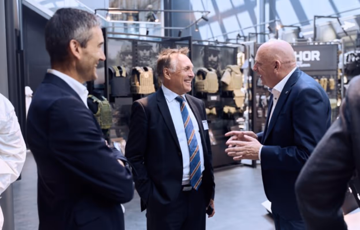 Three men in suits engaged in a conversation in a showroom displaying tactical vests and gear.