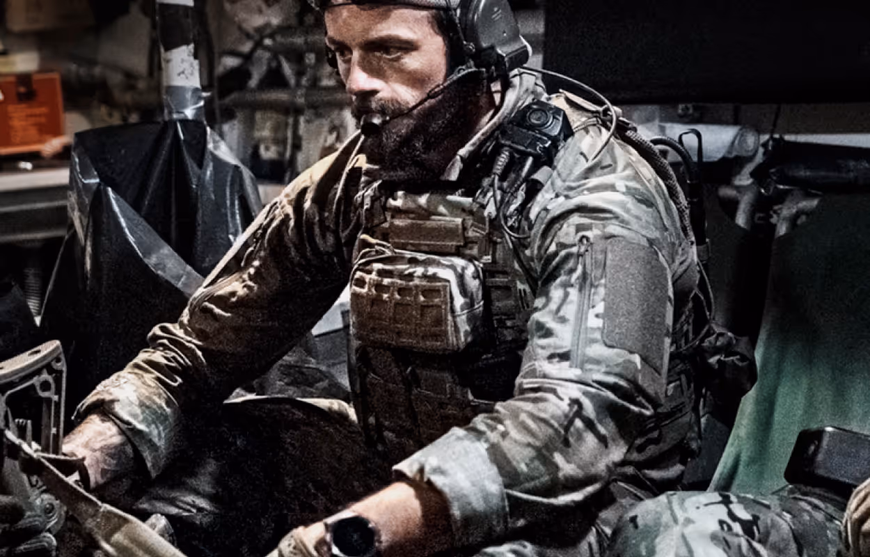 Soldier in camouflage uniform wearing a headset and tactical gear inside a military vehicle.