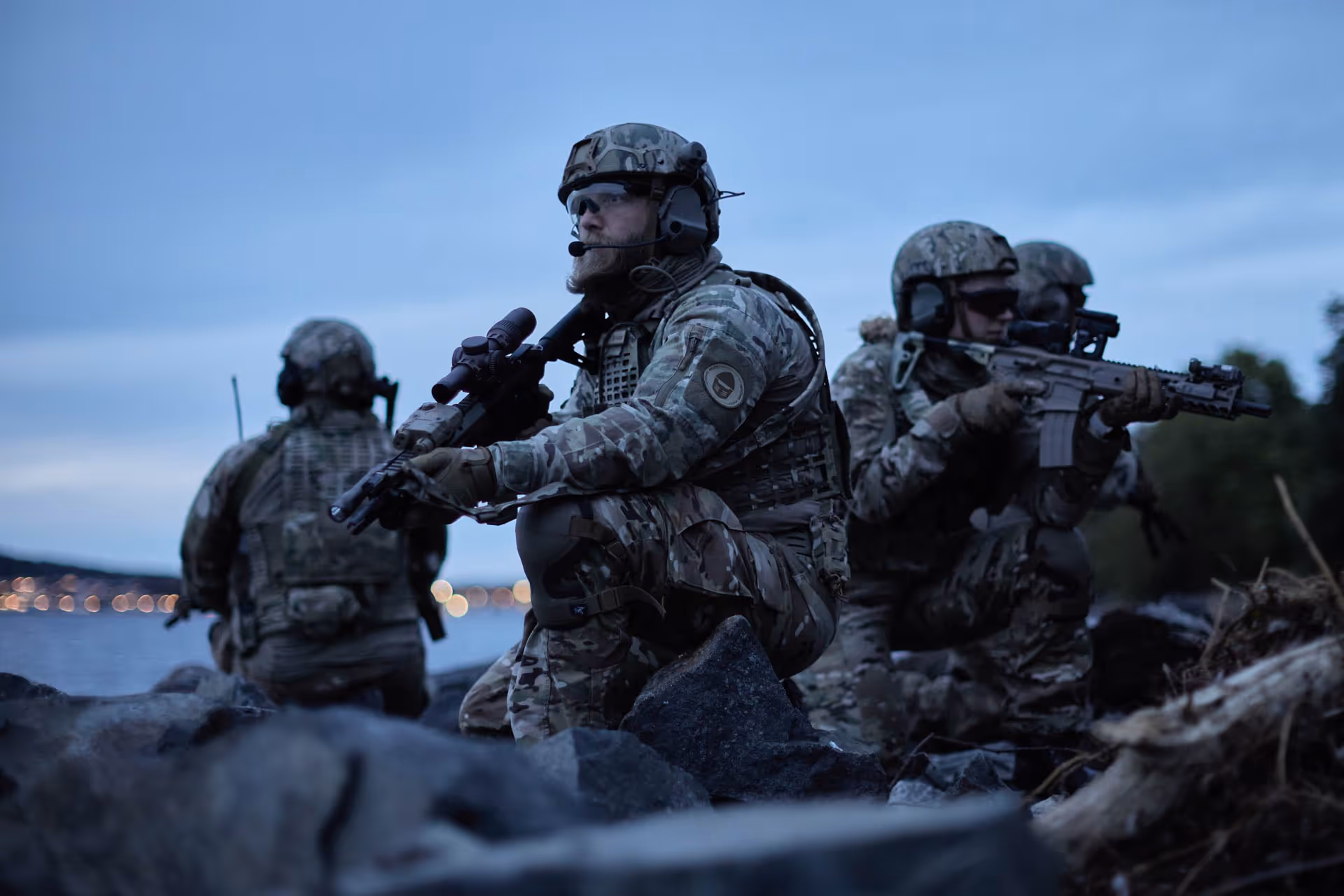 Four soldiers in camouflage gear and helmets are positioned on rocky terrain near water during dusk, holding rifles and appearing alert.