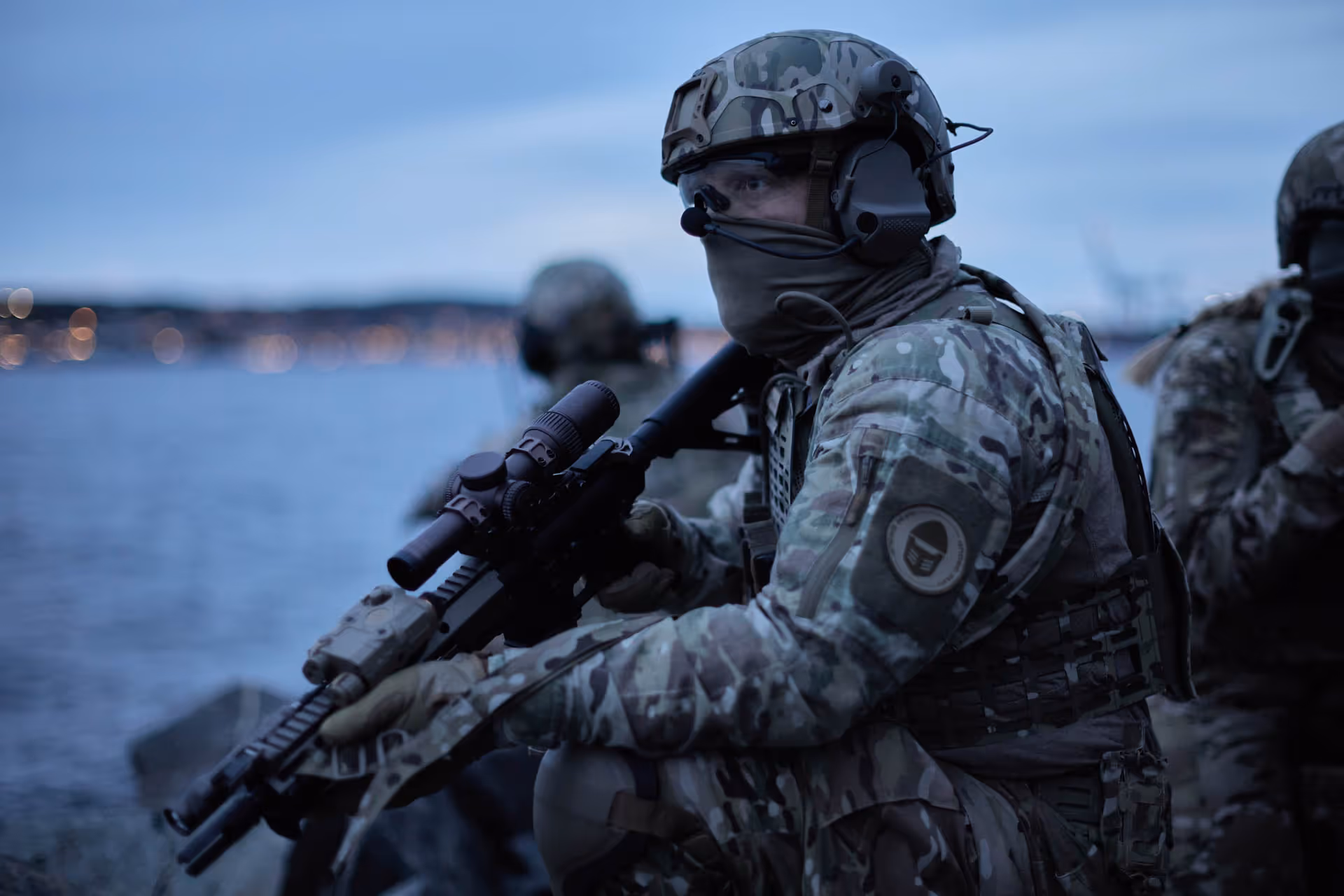 Soldier in camouflage gear and helmet holding a rifle near water at dusk.