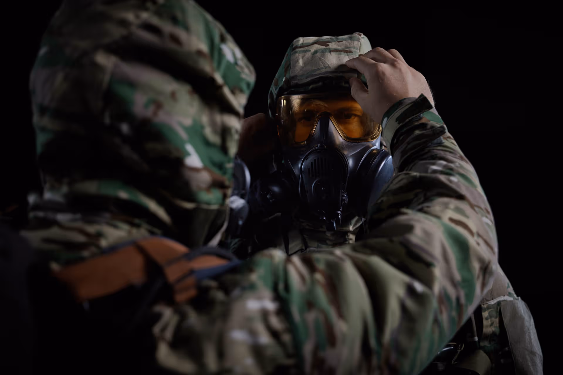 Two soldiers in camouflage uniforms, one adjusting the gas mask of the other against a black background.