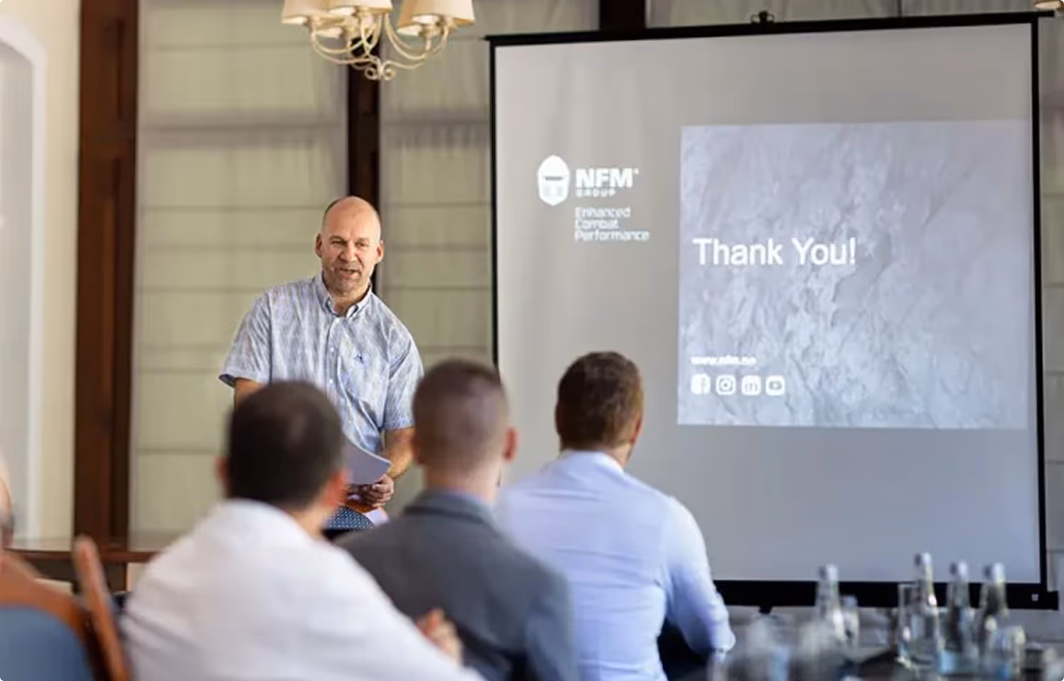 A man in a short-sleeve shirt giving a presentation to an audience seated in front of a screen displaying a 'Thank You!' message and NFM Group logo.