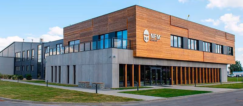 Modern two-story office building with wooden paneling on the upper floor, concrete on the lower floor, and large windows, surrounded by grass and pavement.