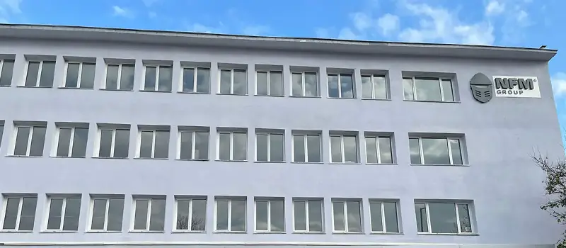 Facade of a white multi-story building with multiple windows and NFM Group logo on the upper right corner under a partly cloudy sky.