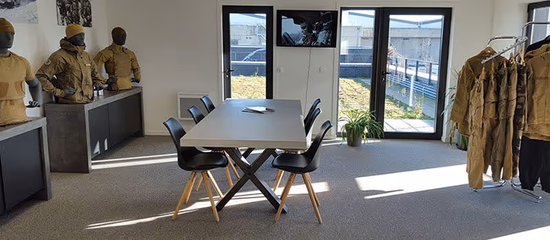 Room with a table surrounded by six black chairs, mannequins dressed in tactical clothing, and a rack of hanging garments near glass doors.