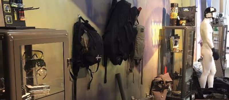 Display case and wall with tactical gear including backpacks, pouches, and a mannequin wearing gloves and a cap in a showroom.