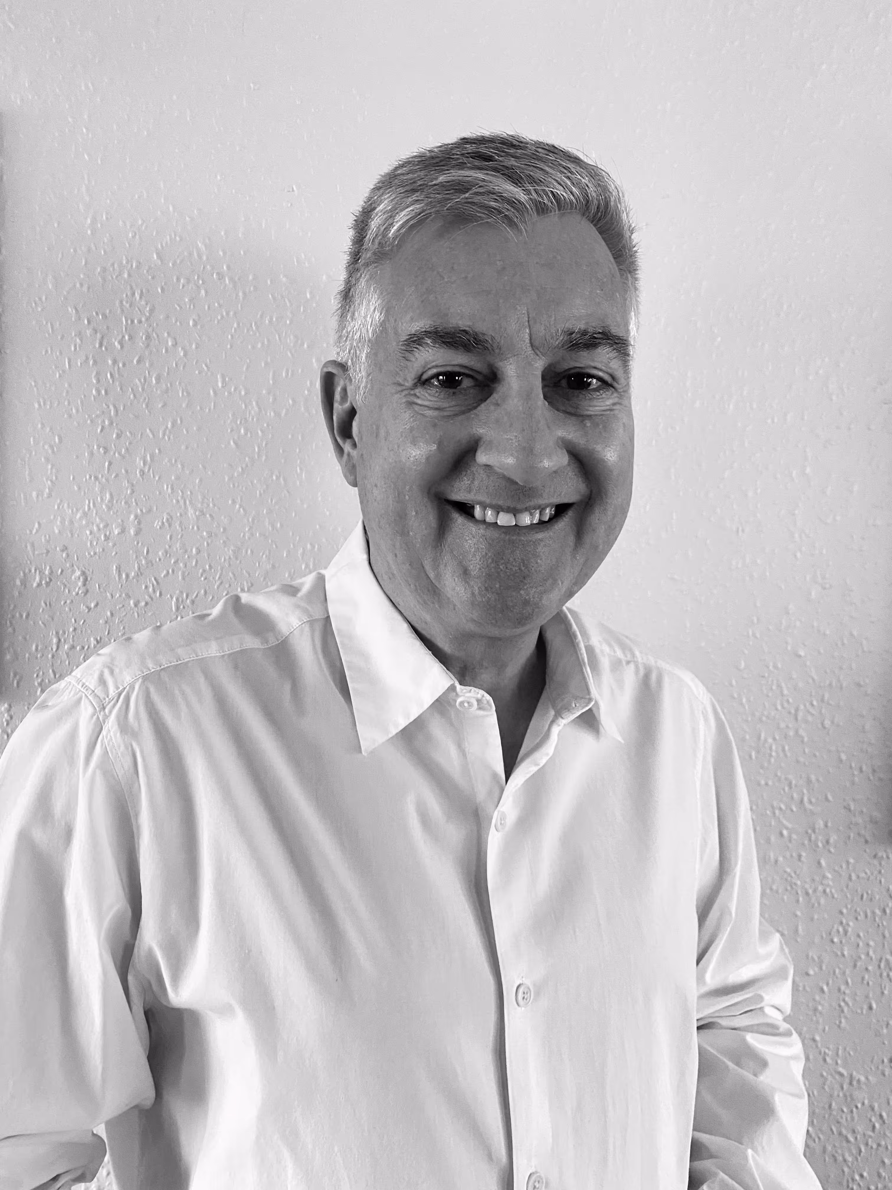 Smiling middle-aged man with gray hair wearing a white button-up shirt against a textured wall.