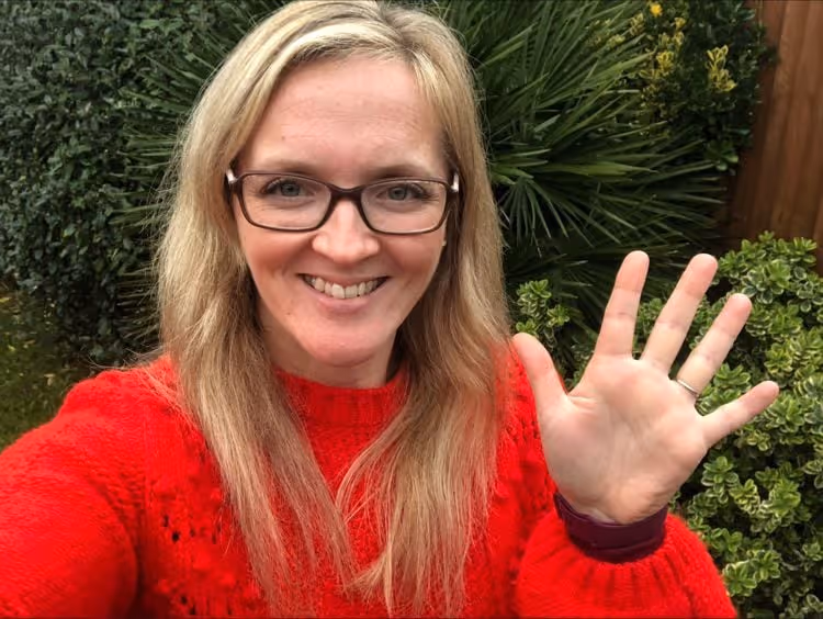 Smiling woman with glasses wearing a red sweater waving her hand outdoors with green bushes in the background.
