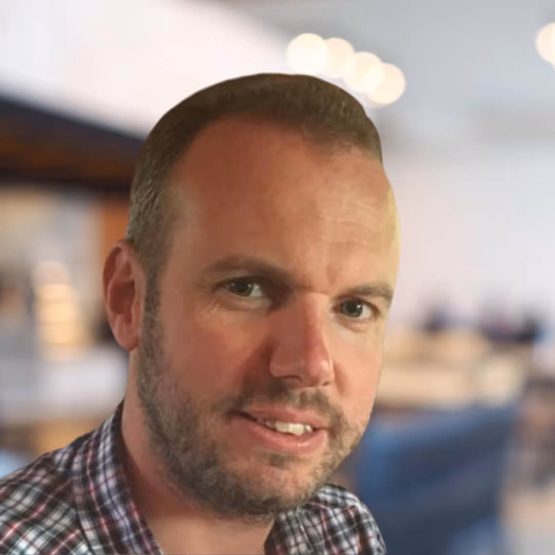 Close-up of a man with short hair and a beard wearing a checkered shirt in a softly lit indoor setting.
