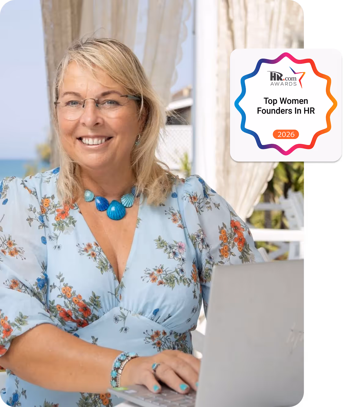Smiling woman with glasses working on a laptop at a seaside outdoor table with a book titled 'Leap Into HR Consulting'.