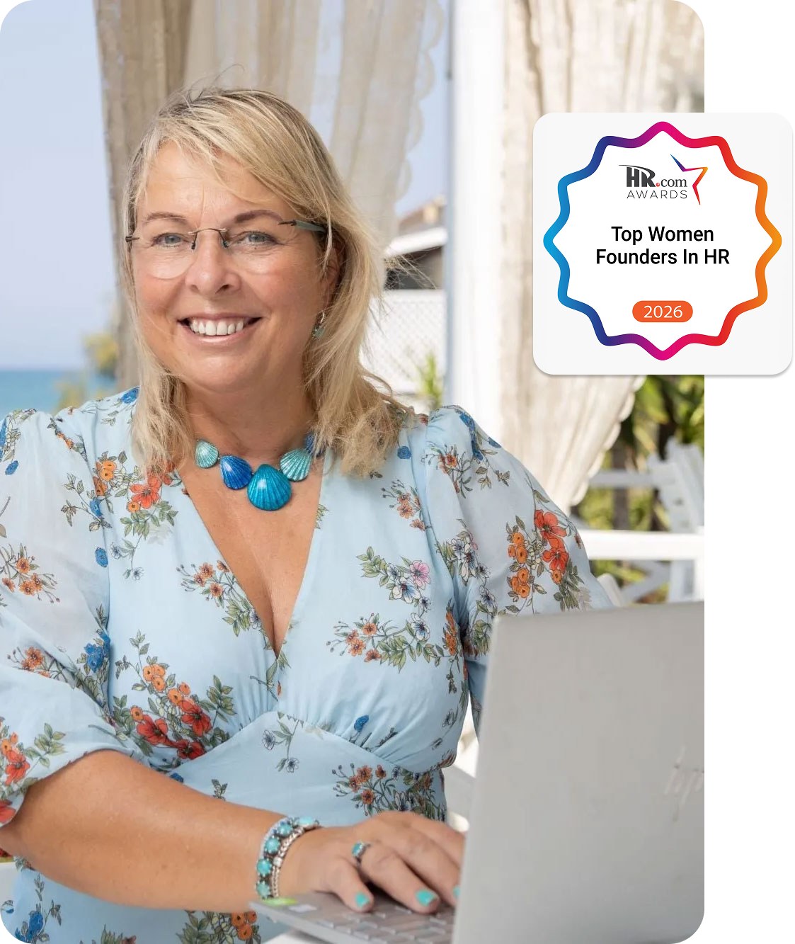 Smiling woman with glasses working on a laptop at a seaside outdoor table with a book titled 'Leap Into HR Consulting'.