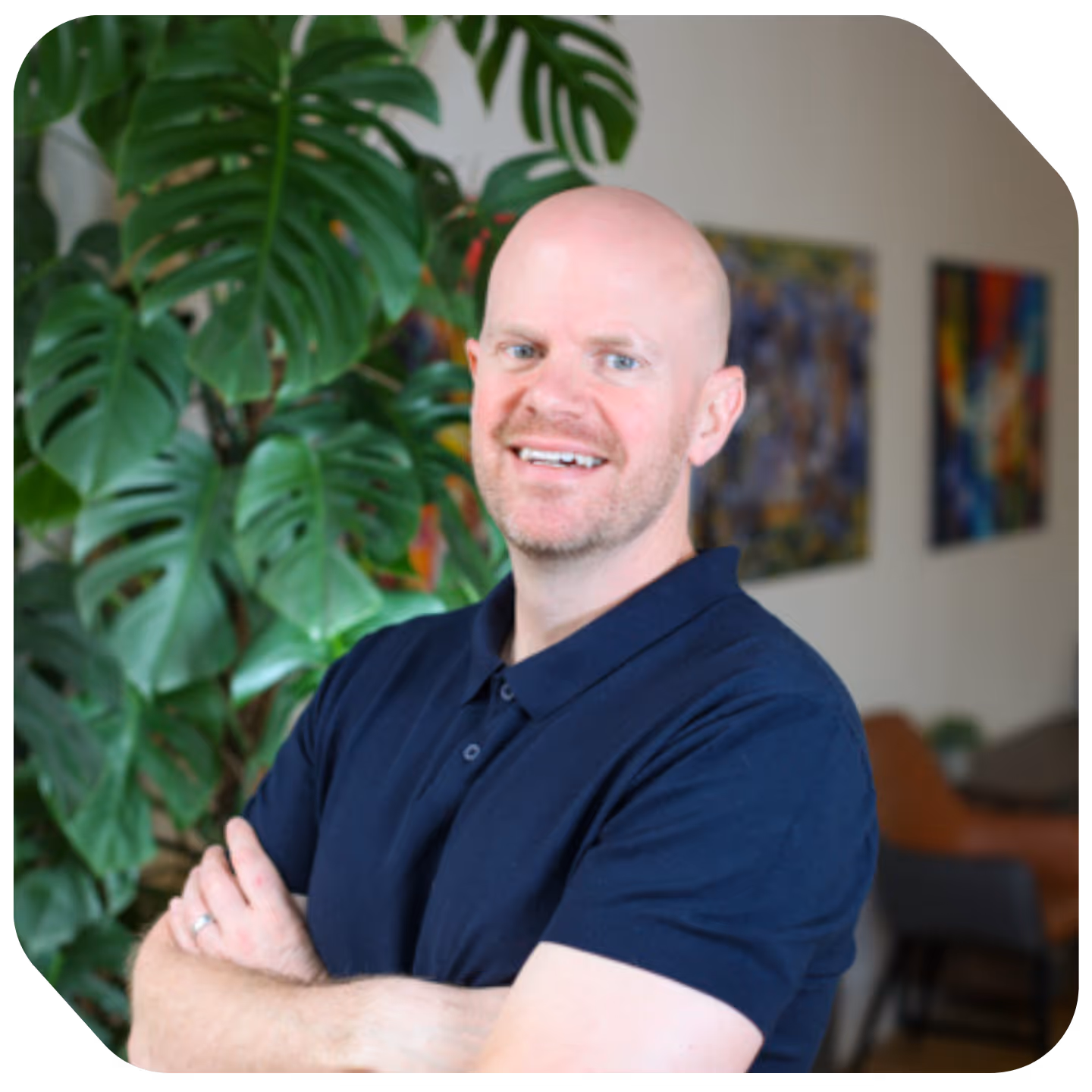 James Bloor in a navy blue polo shirt standing with arms crossed in front of a large green leafy plant indoors.