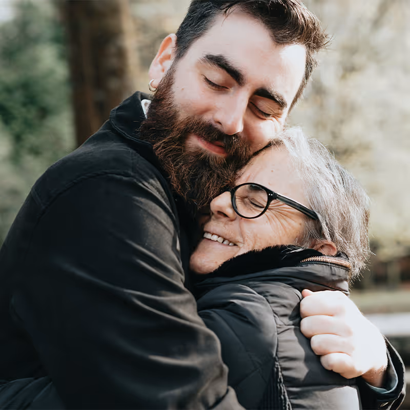 Bearded man hugging and smiling with an older woman wearing glasses outdoors. Clean Slate Clinic clients.