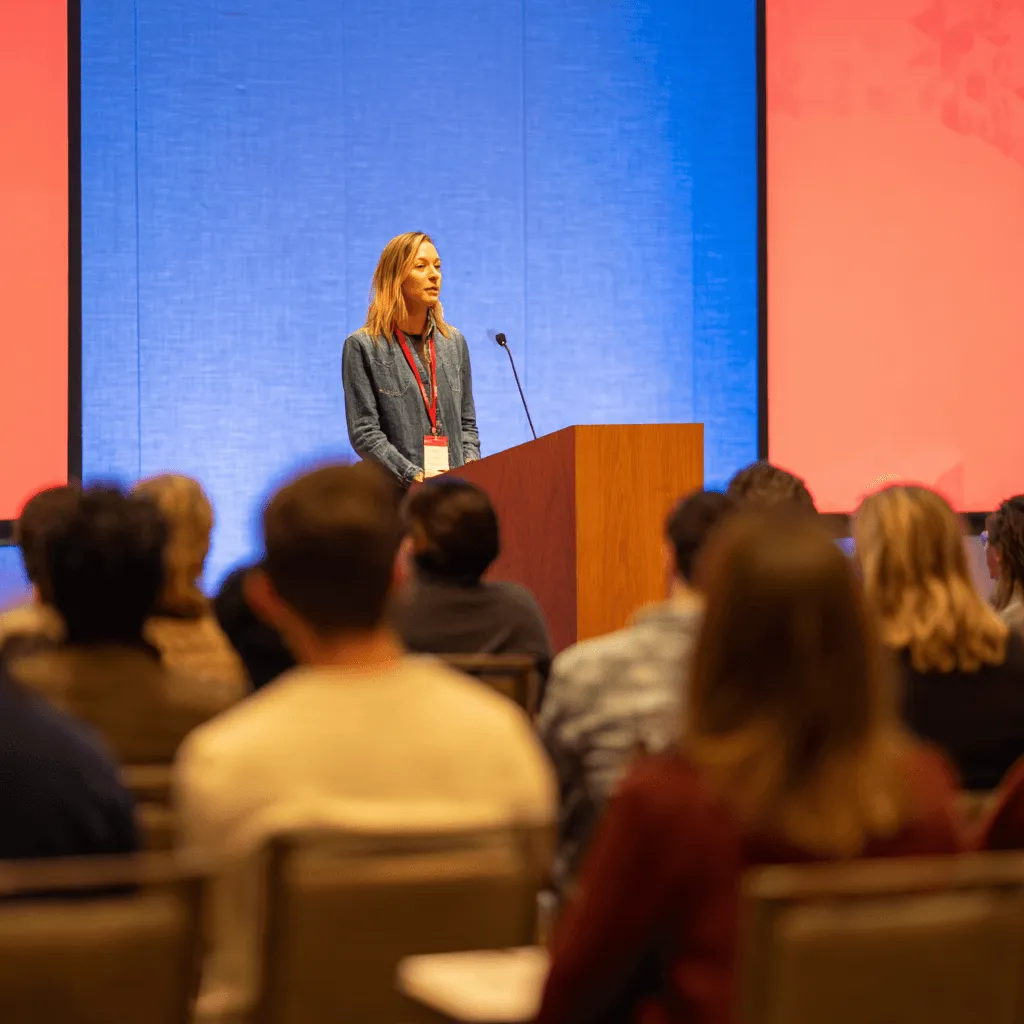 Woman with a red lanyard speaking at a podium in front of an audience with blue and red panels in the background.
