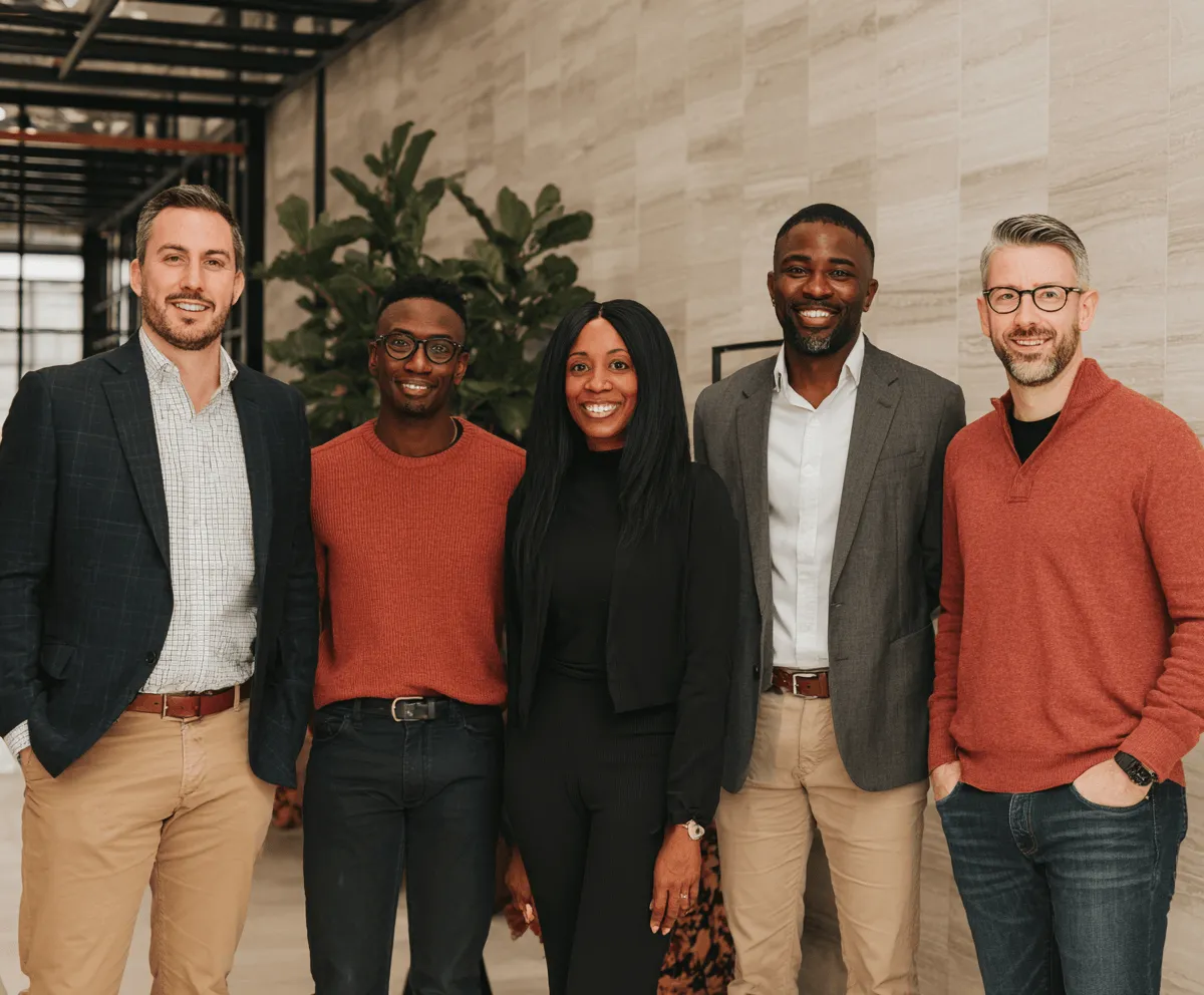 Group of five diverse professionals smiling and standing indoors in business casual attire.
