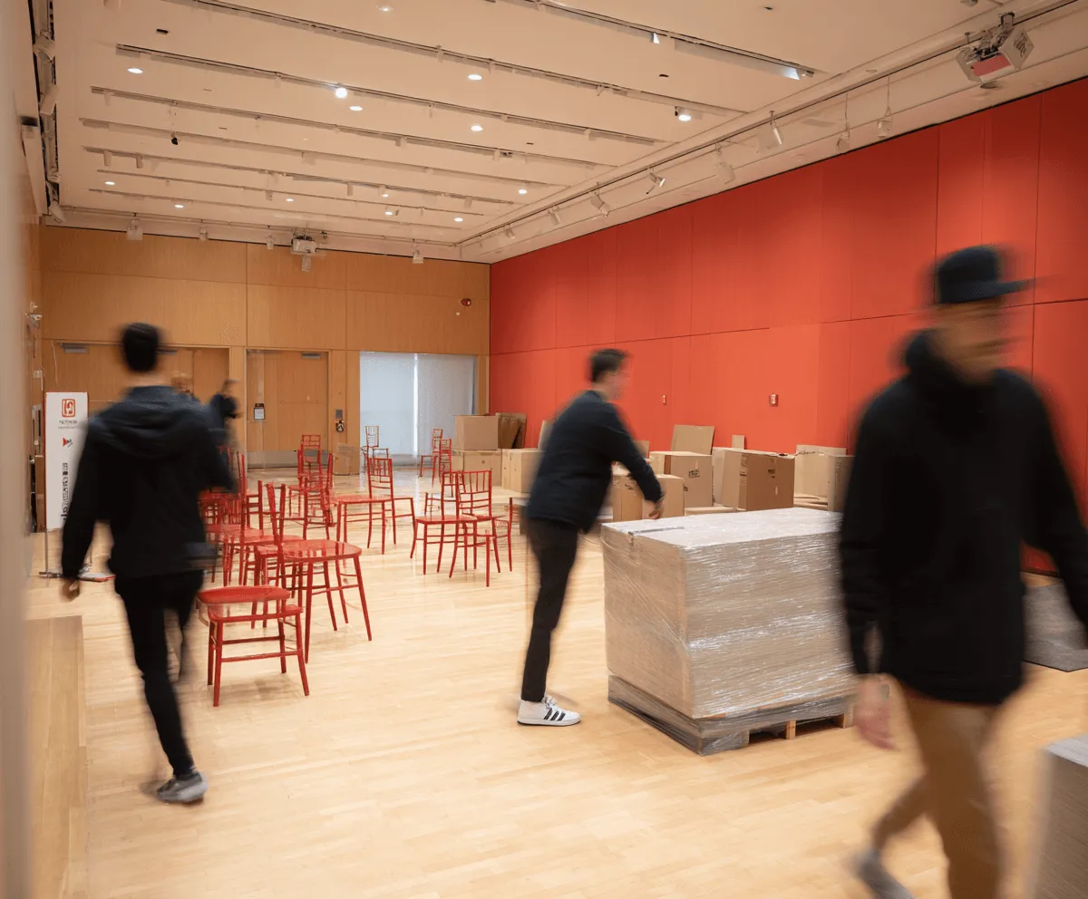 Room with light wood flooring and red wall paneling, several red chairs scattered, and three blurred people moving around near wrapped pallets and cardboard boxes.