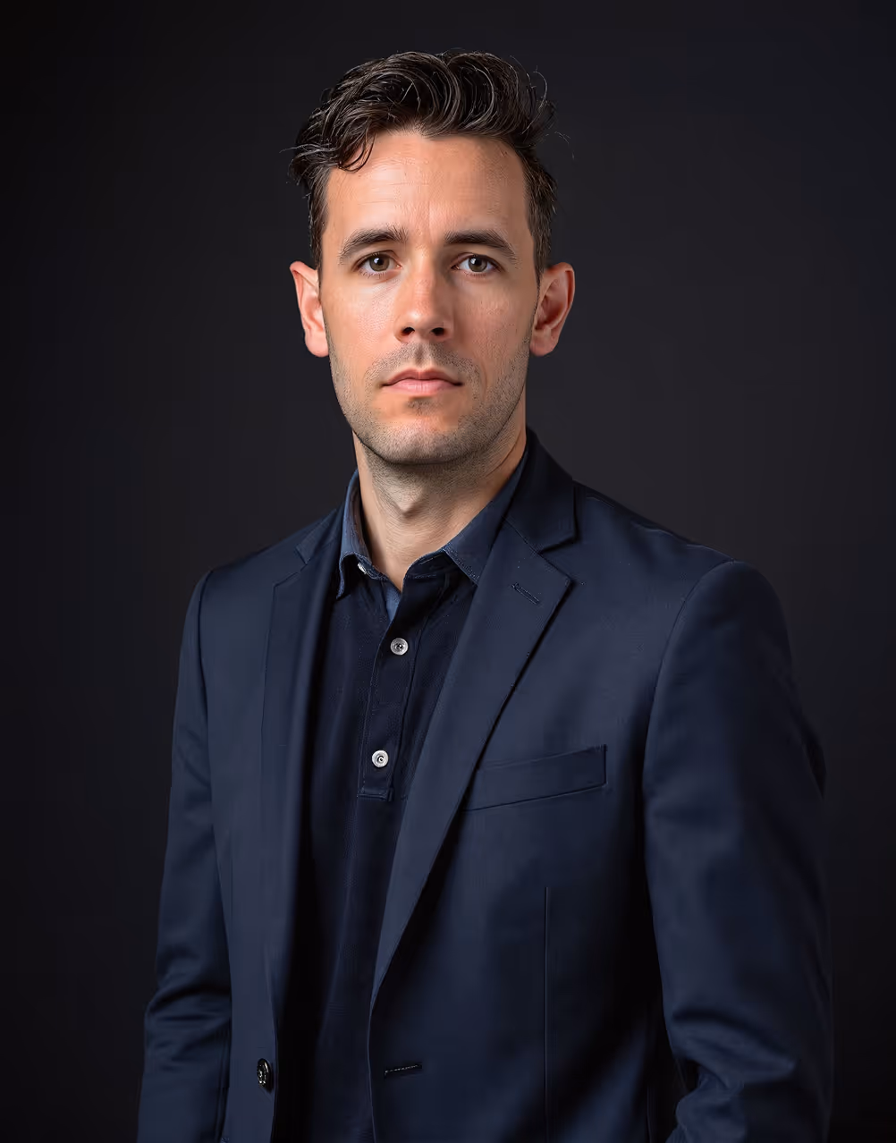 Serious man with dark hair in a navy blazer and shirt against a dark background.